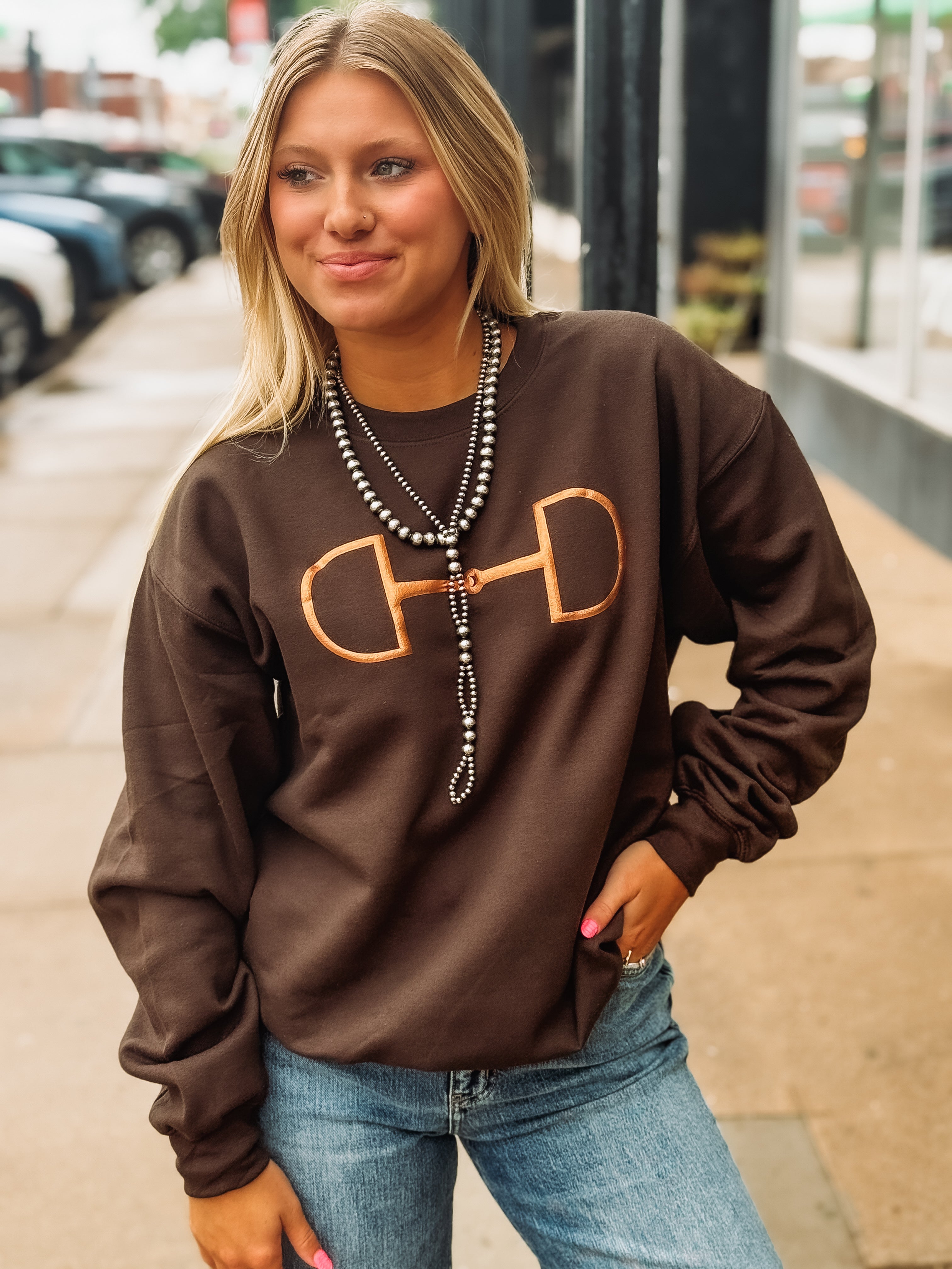 Woman on city street wearing dark brown sweatshirt with metallic bronze horse bit logo