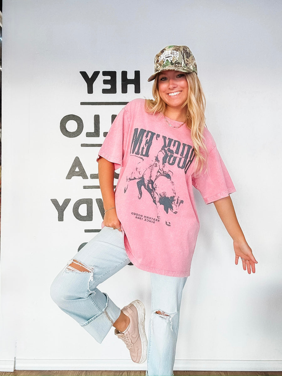 Person wearing a pink graphic t-shirt and light blue jeans with a camouflage cap.
