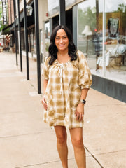 Woman wearing a mustard hued plaid dress standing on a sidewalk with a building in the background.