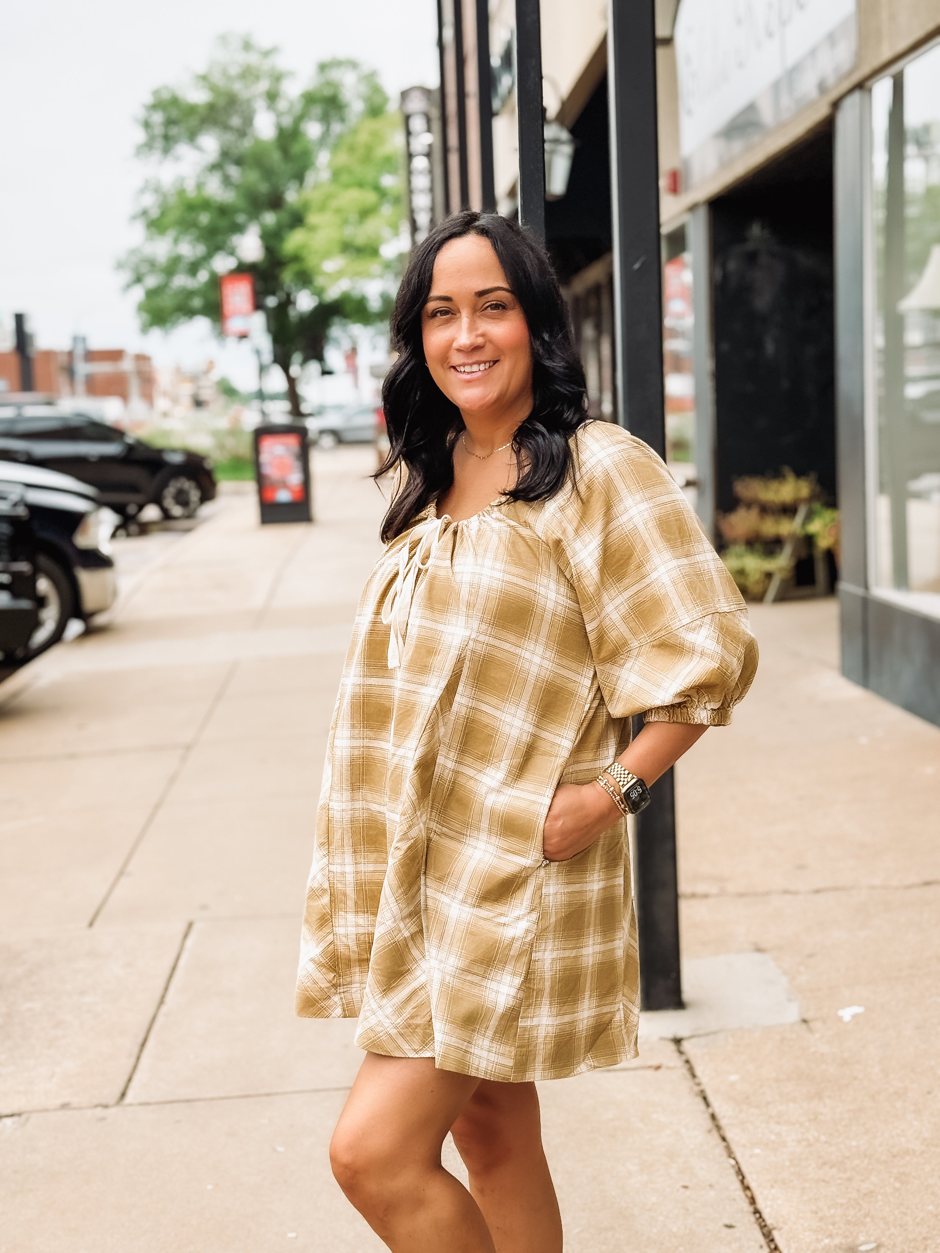 Woman wearing a plaid mustard romper standing on a sidewalk with buildings and cars in the background