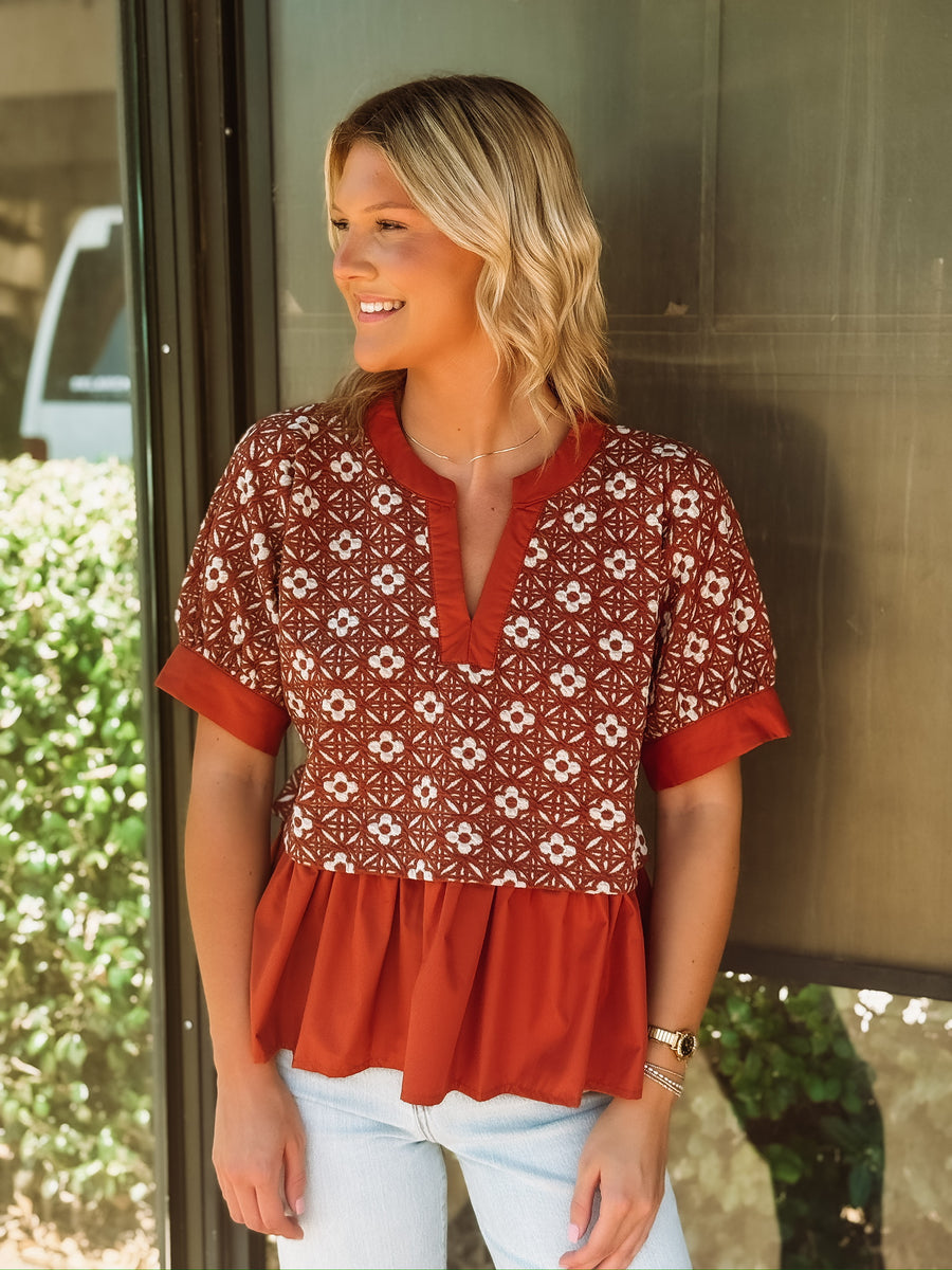 Rust colored short sleeve top with a peplum hem and a mixed media white floral pattern up top.