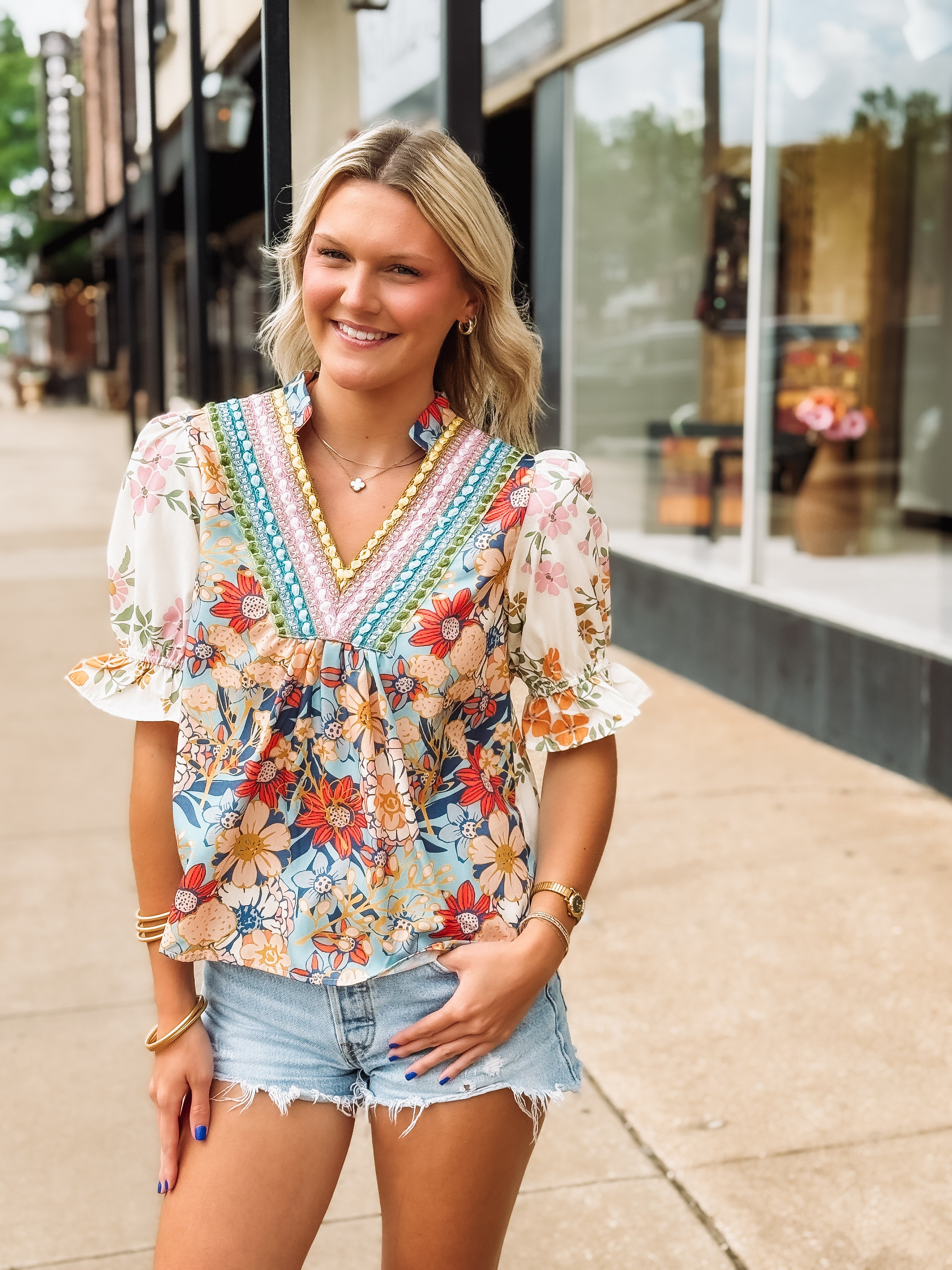Woman wearing a colorful floral blouse and denim shorts standing on a sidewalk.